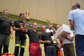Grupa mężczyzn stoi na podwyższeniu podczas wręczania nagród. Dwóch z nich unosi złote puchary, a pozostali trzymają mniejsze trofea i nagrody rzeczowe. Przed nimi stoi mężczyzna w jasnej koszuli.