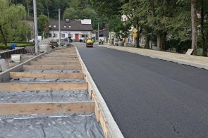 Odcinek drogi z nową nawierzchnią asfaltową podczas prac modernizacyjnych. Po lewej stronie przygotowywany jest chodnik, a po prawej widoczny jest pas zieleni z drzewami. W tle pracują maszyny drogowe i robotnicy.