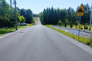 Droga z nawierzchnią asfaltową biegnie przez teren otoczony zielenią i zabudową. Po prawej stronie widoczny jest chodnik oraz znak ostrzegający o spadku drogi wynoszącym 10%.