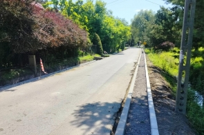 Fragment drogi podczas prac remontowych. Po obu stronach jezdni ułożono nowe krawężniki, a po prawej przygotowano podbudowę pod chodnik. Wzdłuż drogi rosną drzewa i krzewy.