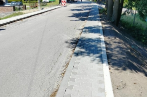 Chodnik z szarej kostki brukowej biegnie wzdłuż drogi otoczonej drzewami. Przy jezdni ustawiono biało-czerwone oznaczenia robót drogowych, a w tle widoczne są maszyny i pracownicy.
