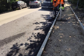 Pracownicy drogowi w pomarańczowych kamizelkach układają nową nawierzchnię asfaltową przy użyciu maszyny drogowej. Obok jezdni przygotowany jest pas pod chodnik, a wzdłuż drogi rosną drzewa.