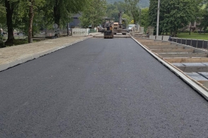 Odcinek drogi z nową nawierzchnią asfaltową podczas prac modernizacyjnych. Po bokach widoczne są nowe krawężniki i przygotowywane fragmenty chodnika, a w tle pracują maszyny drogowe.