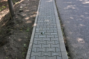 Chodnik z szarej kostki brukowej biegnie wzdłuż drogi, obok zadrzewionego terenu. Przy jezdni ustawiono biało-czerwone oznaczenia robót drogowych.
