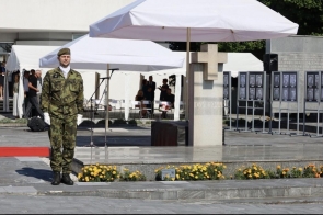 Żołnierz w mundurze stoi na placu przed pomnikiem z krzyżem. W tle widoczne są białe namioty, mikrofony oraz plansze z czarno-białymi portretami.