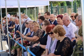 Grupa osób siedzi w kilku rzędach pod białymi namiotami. Część trzyma kwiaty i wieńce, a wśród zgromadzonych widoczne są osoby starsze oraz przedstawiciele służb.