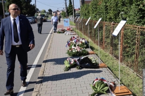 Mężczyzna w garniturze idzie wzdłuż chodnika, przy którym ustawiono tablice informacyjne. Na chodniku leżą wieńce i bukiety kwiatów, a w tle widoczne są kolejne osoby oraz pojazdy.