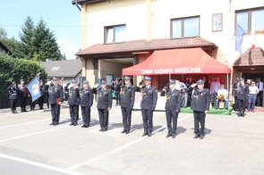 Strażacy w mundurach galowych stoją w szeregu przed remizą i salutują. Po lewej stronie widoczny jest poczet ze sztandarem, a w tle czerwony namiot z napisem „OSP Górki Wielkie” oraz zgromadzone osoby.