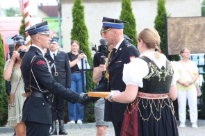 Dwóch strażaków w mundurach galowych stoi naprzeciw siebie podczas wręczania dokumentu lub wyróżnienia. Obok stoi kobieta w stroju regionalnym trzymająca teczkę, a w tle widoczne są osoby obserwujące uroczystość.