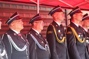 Grupa strażaków w mundurach galowych stoi w szeregu pod czerwonym zadaszeniem. Część ma ozdobne sznury i odznaczenia na mundurach, wszyscy patrzą w tym samym kierunku.