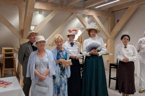 Grupa osób stoi we wnętrzu z drewnianymi belkami konstrukcyjnymi. Kilka kobiet ma stroje stylizowane na dawne, z długimi sukniami i kapeluszami, a jedna trzyma wachlarz.