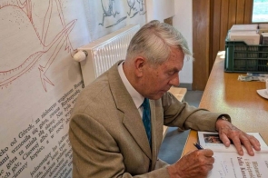 Mężczyzna w garniturze siedzi przy stole i podpisuje książkę „Historia i świętość. Zofia Kossak”. W tle widoczne są ilustracje i teksty umieszczone na ścianie.