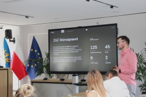 Mężczyzna w różowej koszuli stoi obok dużego ekranu i prowadzi prezentację. Na ekranie widoczny jest slajd dotyczący ZAZ Różnosprawni z danymi liczbowymi. Po lewej stronie stoją flagi a na pierwszym planie siedzą uczestnicy spotkania.