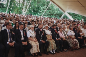 Duża grupa uczestników siedzi na widowni pod zadaszeniem amfiteatru. W pierwszych rzędach widoczne są osoby w strojach regionalnych oraz eleganckich ubraniach, a za nimi licznie zgromadzona publiczność. W tle otaczają ich drzewa i zieleń.