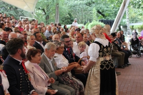 Kobieta w stroju regionalnym rozdaje uczestnikom siedzącym w pierwszym rzędzie drobny poczęstunek z wiklinowego koszyka. Wokół zgromadzona jest liczna publiczność, w tym osoby w strojach regionalnych i eleganckich ubraniach. W tle widoczna jest zadaszona widownia otoczona zielenią.
