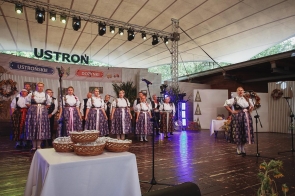 Grupa osób w strojach regionalnych występuje na scenie podczas dożynek. Po prawej stronie kobieta śpiewa do mikrofonu, a za nią stoi większy zespół. Na pierwszym planie znajduje się stół z wiklinowymi koszami.
