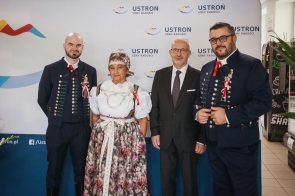 Cztery osoby pozują do zdjęcia na tle ścianki promocyjnej Ustronia. Dwóch mężczyzn ma na sobie stroje regionalne, kobieta również ubrana jest w strój regionalny, a stojący obok mężczyzna ma ciemny garnitur.