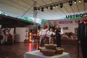 Kobieta w stroju regionalnym przemawia do mikrofonu na scenie. Za nią stoją inni uczestnicy w strojach regionalnych, a z boku widoczni są muzycy. Na pierwszym planie ustawiono kosze z pieczywem.