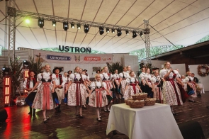 Zespół w strojach regionalnych występuje na scenie podczas dożynek. Z przodu ustawiono stół z koszami pieczywa, a jedna z kobiet śpiewa do mikrofonu.