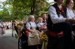 Uczestnicy dożynkowego korowodu jadą bryczką udekorowaną kwiatami i kłosami zbóż. Kobieta i mężczyzna mają na sobie stroje regionalne, a między nimi siedzi mała dziewczynka. W tle widoczni są kolejni uczestnicy korowodu oraz licznie zgromadzeni widzowie.