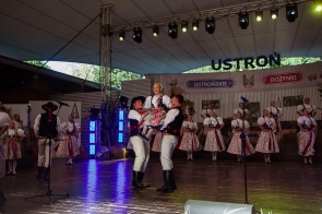 Dwóch mężczyzn w strojach regionalnych unosi dziewczynkę podczas występu na scenie. W tle stoją pozostali członkowie zespołu w regionalnych strojach, a nad sceną widoczne są dekoracje z napisami „Ustroń” i „Ustrońskie Dożynki”.