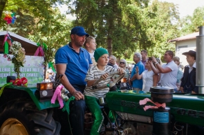 Mężczyzna siedzi na zielonym ciągniku razem z dwójką chłopców podczas dożynkowego korowodu. Jeden z chłopców trzyma kierownicę, a pojazd ozdobiony jest kolorowymi wstążkami i kwiatami. W tle stoją licznie zgromadzeni widzowie.
