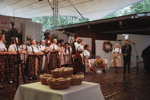Na scenie stoją uczestnicy uroczystości w strojach regionalnych. Kobiety po lewej stronie trzymają bochenki chleba, a na pierwszym planie widoczne są wiklinowe kosze z pieczywem. Po prawej stronie stoją kolejne osoby w strojach regionalnych, a scenę zdobi wieniec i kompozycje kwiatowe.