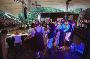 Grupa osób w strojach regionalnych stoi na scenie podczas występu. Uczestnicy ustawieni są w kręgu i trzymają się za ręce, a po lewej stronie widoczna jest licznie zgromadzona publiczność. Na scenie znajdują się także stoły z pieczywem i dekoracjami dożynkowymi.