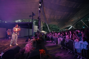 Wokalista występuje na scenie przed licznie zgromadzoną publicznością. Śpiewa do mikrofonu, a w tle widoczny jest gitarzysta. Widownia stoi za barierkami pod zadaszeniem amfiteatru, a całość oświetlona jest fioletowym światłem scenicznym.