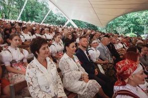 Licznie zgromadzona publiczność siedzi na widowni amfiteatru pod zadaszeniem. W pierwszych rzędach widoczne są osoby w strojach regionalnych oraz eleganckich ubraniach, a dalej kolejne rzędy uczestników wydarzenia.