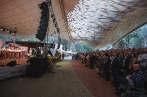 Licznie zgromadzona publiczność stoi pod zadaszeniem amfiteatru i obserwuje uroczystość. Po lewej stronie na scenie widoczne są osoby w strojach regionalnych, a wśród widowni także przedstawiciele służb i zaproszeni goście. Nad całością rozciąga się duże zadaszenie z dekoracyjnym oświetleniem.