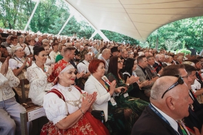 Licznie zgromadzona publiczność siedzi na widowni amfiteatru pod zadaszeniem. W pierwszych rzędach widoczne są osoby w strojach regionalnych oraz eleganckich ubraniach, a wiele osób bije brawo.