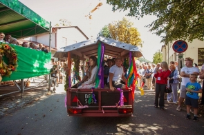 Udekorowana przyczepa jedzie w dożynkowym korowodzie. W środku siedzi kilka osób, a pojazd ozdobiony jest kolorowymi wstążkami i zielenią. Po obu stronach trasy stoją licznie zgromadzeni widzowie, a po lewej stronie widoczna jest zadaszona trybuna.