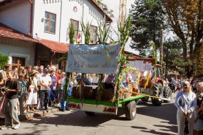 Udekorowana przyczepa ciągnięta przez traktor jedzie w dożynkowym korowodzie. Przyczepę ozdobiono kukurydzą, słonecznikami, kolorowymi wstążkami i belami słomy, a z tyłu umieszczono napis „Kołocz, chleb i dobre słowo – z KGW zawsze odlotowo”. Po obu stronach ulicy stoją licznie zgromadzeni widzowie.