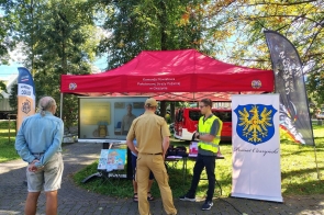 Stoisko informacyjne w parku pod czerwonym namiotem. Przed ekranem z prezentacją stoją uczestnicy oraz przedstawiciele służb, a obok znajduje się baner z herbem Powiatu Cieszyńskiego i flagi promocyjne programu OLiOC oraz Ustronia.