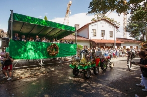 Udekorowany ciągnik z przyczepą jedzie w dożynkowym korowodzie przed licznie zgromadzonymi widzami. Przyczepa ozdobiona jest zielenią, kwiatami i elementami nawiązującymi do rolnictwa. Po lewej stronie znajduje się zadaszona trybuna z uczestnikami uroczystości, a w tle widoczny jest budynek.