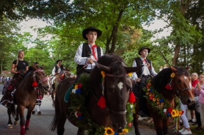 Grupa osób w strojach regionalnych jedzie konno w dożynkowym korowodzie. Konie są udekorowane kolorowymi kwiatami, zielonymi girlandami i wstążkami. Po obu stronach trasy stoją licznie zgromadzeni widzowie.