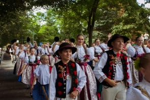 Grupa dzieci i młodzieży w strojach regionalnych uczestniczy w korowodzie, idąc alejką wśród drzew. Na pierwszym planie widoczni są chłopcy w kapeluszach i ozdobnych kamizelkach, a za nimi dziewczęta w białych bluzkach i długich spódnicach. Część uczestników macha w stronę zgromadzonych osób.