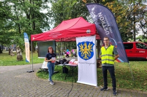 Kobieta i mężczyzna stoją obok stoiska informacyjnego w parku. Między nimi znajduje się baner z herbem Powiatu Cieszyńskiego, a w tle czerwony namiot, ekran z prezentacją oraz stół z materiałami informacyjnymi. Po prawej stronie widoczna jest flaga promująca Ustroń.