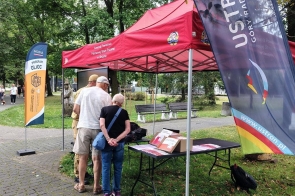 Czerwony namiot stoi w parku przy alejce. Pod namiotem ustawiono stół z materiałami informacyjnymi, przy którym stoją trzy osoby. Obok znajdują się flagi.