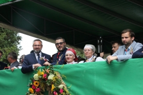 Grupa osób stoi na zadaszonej trybunie i obserwuje wydarzenie. Wśród nich są osoby w strojach regionalnych oraz eleganckich ubraniach. Z przodu trybuny widoczna jest zielona osłona ozdobiona dużym wieńcem z kwiatów, kłosów i zieleni.