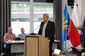 Mężczyzna przemawia przy mównicy w sali sesyjnej. Po prawej stronie widoczne są flagi Powiatu Cieszyńskiego, Polski i Unii Europejskiej, a w tle duże okna sali.