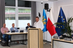 Mężczyzna przemawia przy mównicy w sali sesyjnej. Po prawej stronie widoczne są flagi Powiatu Cieszyńskiego, Polski i Unii Europejskiej, a w tle duże okna sali.