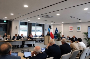 Przy mównicy stoi kobieta przemawiająca do zgromadzonych, a uczestnicy siedzą przy stołach wyposażonych w mikrofony, tablety i tabliczki z nazwiskami. W tle widoczne są flagi.