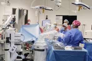 Wokół pacjenta pracuje kilku członków zespołu w strojach ochronnych, a obok widoczne są ramiona robota, aparatura medyczna oraz narzędzia chirurgiczne. Nad stołem operacyjnym znajdują się lampy zabiegowe.