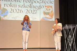 Dwie kobiety stoją na scenie podczas uroczystości rozpoczęcia roku szkolnego. Jedna z nich przemawia do mikrofonu, a druga trzyma bukiet kwiatów. W tle wyświetlana jest prezentacja, a obok widoczna jest mównica.