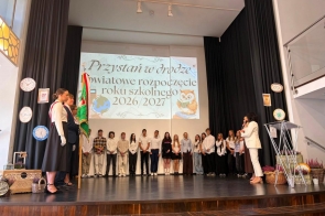 Grupa uczniów stoi w szeregu na scenie podczas uroczystości rozpoczęcia roku szkolnego. Po lewej stronie widoczny jest poczet sztandarowy z biało-czerwonymi szarfami, a po prawej kobieta przemawia do mikrofonu. W tle wyświetlana jest prezentacja, a po bokach sceny znajdują się zegary oraz dodatkowe dekoracje.