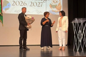 Trzy osoby stoją na scenie podczas uroczystości. Kobieta pośrodku przemawia do mikrofonu, mężczyzna obok trzyma bukiet kwiatów, a druga kobieta stoi przy mównicy. W tle wyświetlana jest prezentacja.