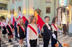 Młodzież stoi w poczcie sztandarowym we wnętrzu kościoła podczas uroczystości. Uczestnicy mają biało-czerwone szarfy i białe rękawiczki, a w tle widoczne są kolejne poczty sztandarowe oraz zgromadzone osoby.