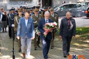 Trzech mężczyzn idzie w kierunku miejsca uroczystości, a jeden z nich niesie wiązankę biało-czerwonych kwiatów. Za nimi maszerują przedstawiciele służb mundurowych, część z nich również trzyma kwiaty. W tle widoczni są uczestnicy wydarzenia oraz zaparkowane samochody.